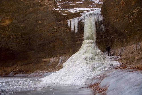 A frozen waterfall in a rocky canyon, with a person standing nearby, surrounded by ice and snow.