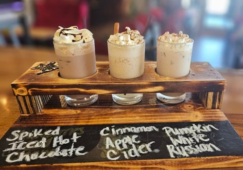 Three festive drinks on a wooden tray: Spiked Treat Hot Chocolate, Cinnamon Apple Cider, and Pumpkin White Russian.