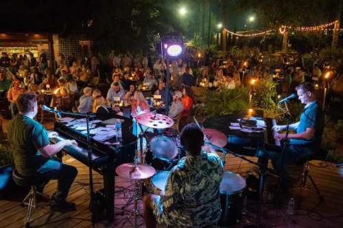 A lively outdoor music performance with a band playing to an engaged audience seated at tables under string lights.