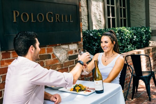A couple toasts with wine glasses at an outdoor restaurant table, enjoying a meal together.