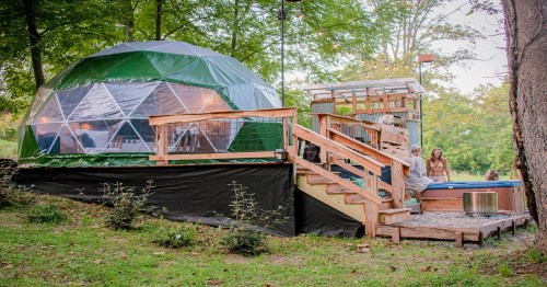 A geodesic dome structure with a wooden deck and hot tub, surrounded by trees in a serene outdoor setting.