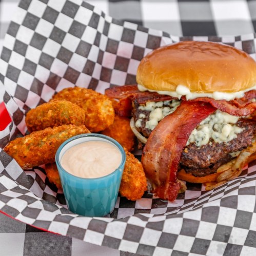 A delicious burger topped with bacon and blue cheese, served with crispy onion rings and a dipping sauce on a checkered tablecloth.