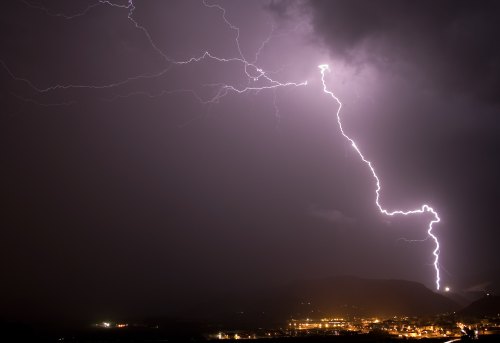 A dramatic night sky illuminated by bright lightning strikes over a dark landscape and distant city lights.
