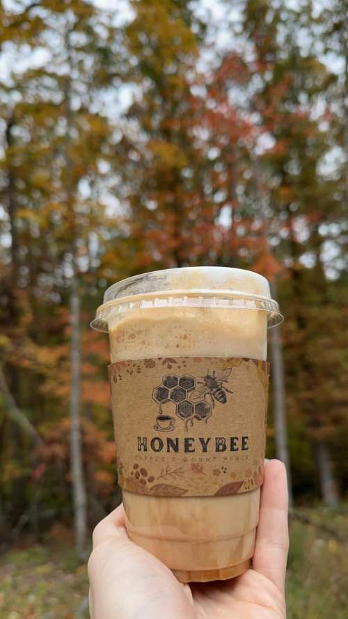 A hand holding a cup of coffee with a "Honeybee" label, set against a backdrop of autumn trees.