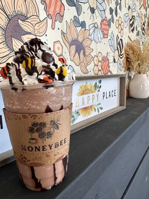 A chocolate milkshake topped with whipped cream, chocolate drizzle, and candy, beside a floral wall and decorative sign.