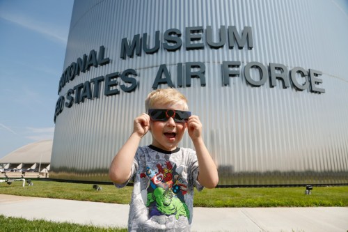 A child wearing eclipse glasses smiles in front of the National Museum of the United States Air Force.