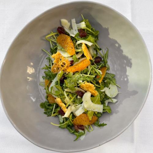 A fresh salad with greens, orange segments, fennel, and raisins in a gray bowl on a white tablecloth.