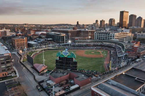 Aerial view of a bustling baseball stadium surrounded by a city skyline at sunset.