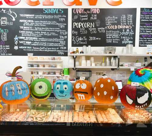 A display of creatively painted pumpkins in front of a dessert counter, featuring various colorful designs.