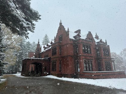 A large, red brick mansion covered in snow, surrounded by trees, with snowflakes gently falling.