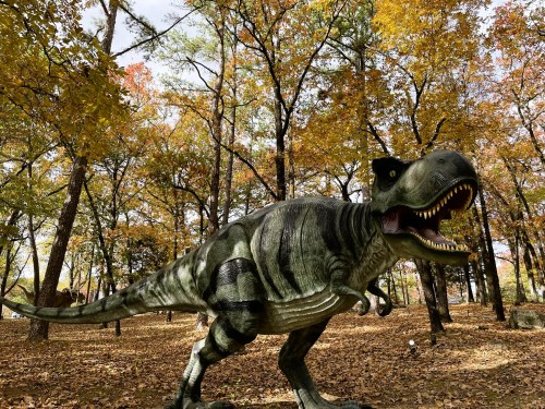 A lifelike dinosaur statue stands among colorful autumn trees in a forest setting.