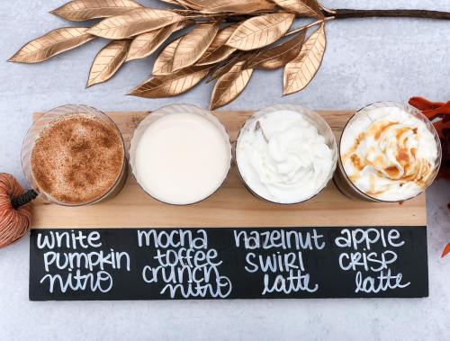 Four coffee drinks in clear cups on a wooden board, labeled: White Pumpkin Nitro, Mocha Toffee Crunch Nitro, Hazelnut Swirl Latte, Apple Crisp Latte.