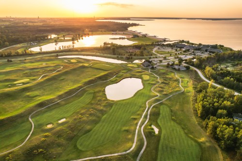 Aerial view of a lush golf course at sunset, featuring ponds and a serene lake in the background.