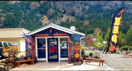A cozy BBQ restaurant with a welcoming sign, surrounded by trees and mountains, featuring outdoor seating and an American flag.