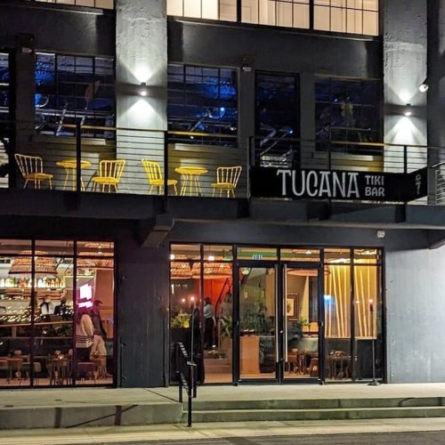 Exterior view of Tucana Tiki Bar at night, featuring large windows, outdoor seating, and a vibrant atmosphere.