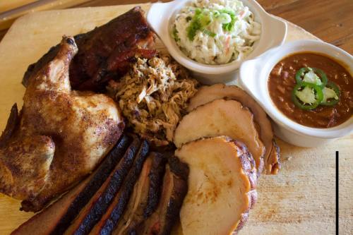 A wooden platter featuring smoked meats: chicken, pulled pork, brisket, and sides of coleslaw and beans.
