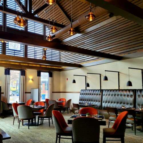 Modern restaurant interior with wooden beams, pendant lights, and cozy seating arrangements. Large windows let in natural light.