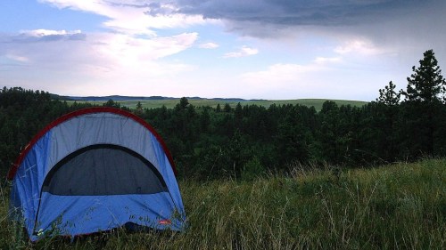 A blue tent set on a grassy hill, surrounded by trees, with a cloudy sky and distant rolling hills in the background.