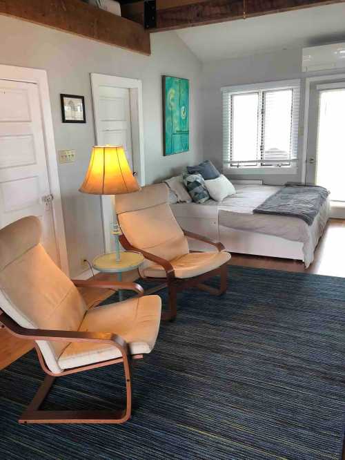 Cozy room with two armchairs, a lamp, a bed, and large windows, featuring light colors and a blue rug.