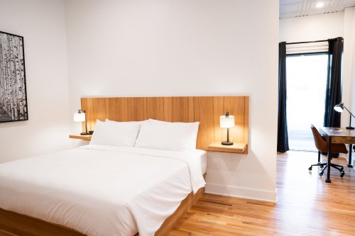 A modern bedroom featuring a queen bed, wooden headboard, bedside lamps, and a workspace by a window.