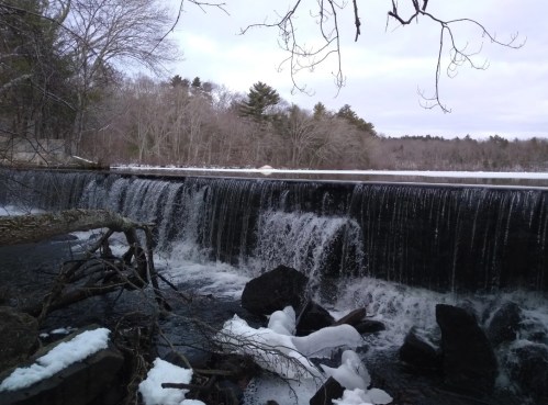 A serene waterfall cascades over a rocky ledge, surrounded by trees and snow, under a cloudy sky.