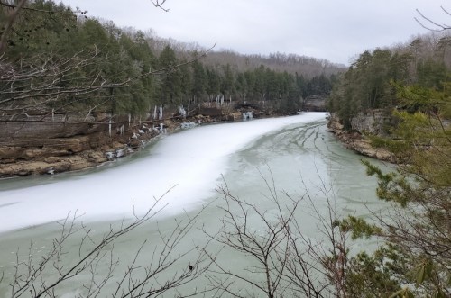 A serene river partially frozen, surrounded by trees and rocky cliffs under a cloudy sky.