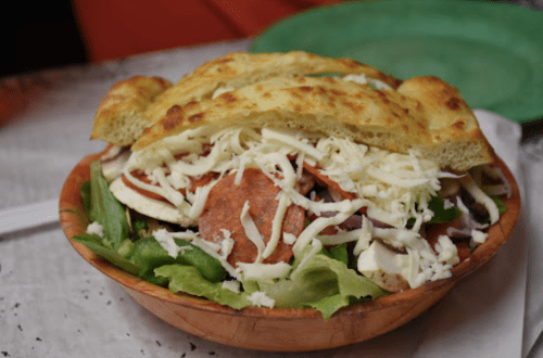 A wooden bowl filled with salad, topped with pepperoni, cheese, and a large piece of bread.