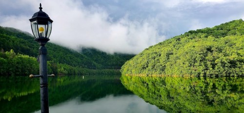 A serene lake surrounded by lush green hills, with a lamppost in the foreground and misty clouds above.