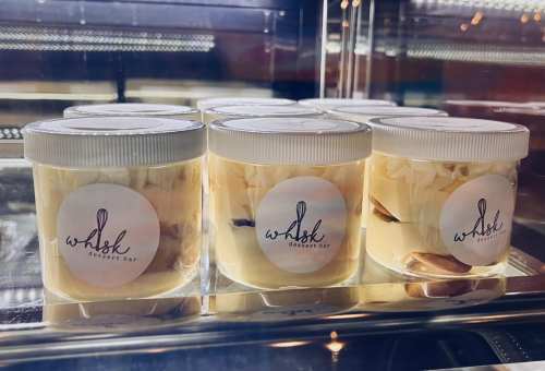 Clear containers of layered desserts with a logo for "Whisk Dessert Bar" displayed in a glass case.