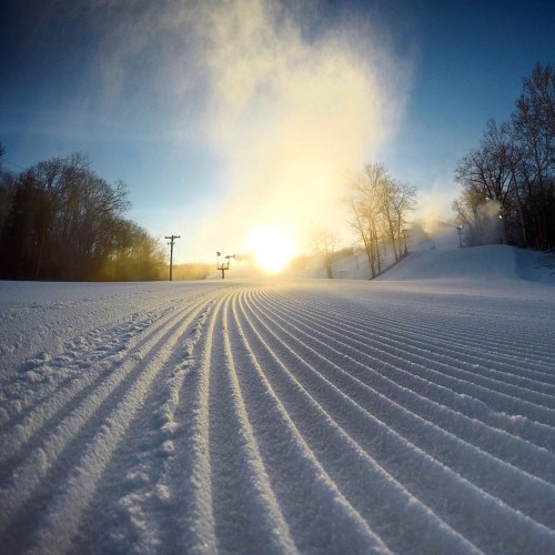 A snowy landscape with groomed ski trails, trees in the background, and a bright sun rising in the sky.