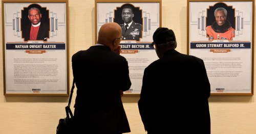 Two people observe framed portraits and biographies of notable individuals on a gallery wall.