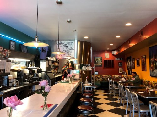 A colorful diner interior with a counter, flowers on tables, and a cozy seating area. A customer is seated in the background.