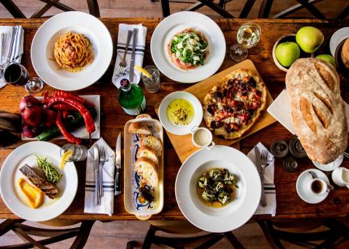 A table set with various dishes, including pasta, salad, pizza, bread, and fresh produce, creating a vibrant dining scene.