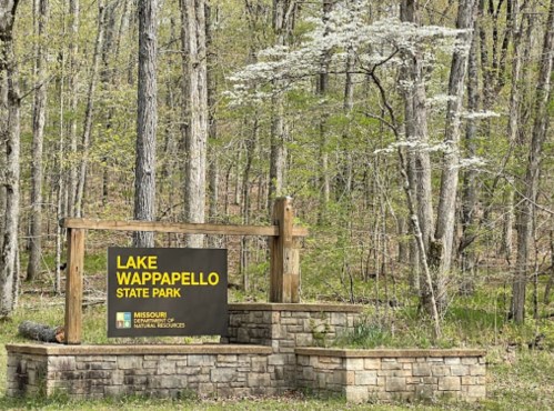 Sign for Lake Wappapello State Park surrounded by trees and greenery in a natural setting.
