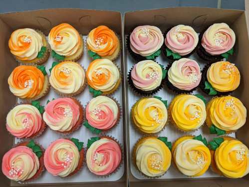 A box of beautifully decorated cupcakes with rose-shaped frosting in orange, pink, and yellow colors, garnished with green leaves.