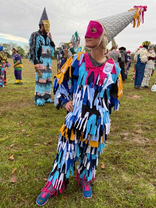 A person in a colorful, fringed costume poses confidently, wearing a cone-shaped hat at a festive outdoor event.