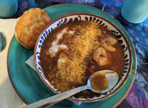 A bowl of shrimp stew with cheese and rice, accompanied by a biscuit on a colorful table.
