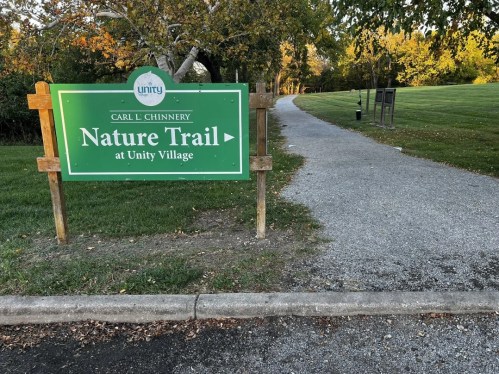 Sign for "Carl L. Chinnery Nature Trail at Unity Village" with a gravel path leading into a green area.