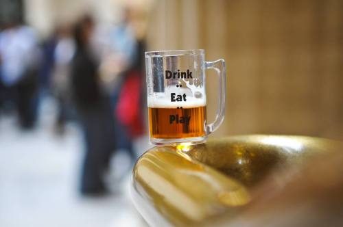 A clear mug of beer sits on a golden surface, with the words "Drink Eat Play" printed on the side.