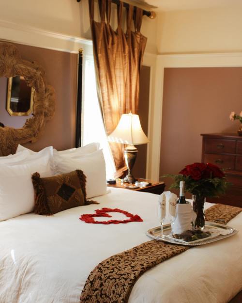 A cozy bedroom with a neatly made bed, decorative pillows, a tray with wine and roses, and elegant curtains.