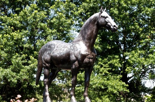 A large bronze statue of a horse stands amidst lush green trees.