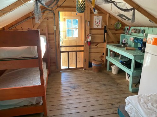 Cozy cabin interior with bunk beds, a kitchenette, and a door leading outside, featuring wooden walls and floors.
