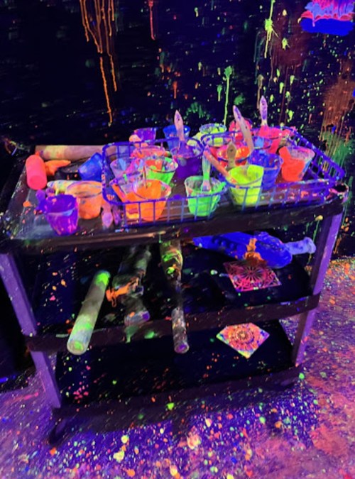 A cart filled with vibrant, glowing paint and brushes, surrounded by splashes of neon colors on a dark background.