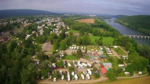 Aerial view of a campground near a river, surrounded by greenery and small buildings in a rural landscape.