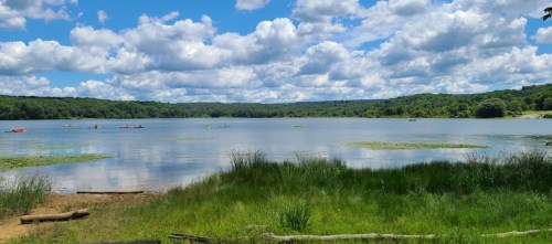 A serene lake surrounded by lush greenery and fluffy clouds, with kayakers enjoying the water on a sunny day.