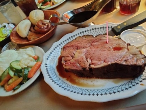 A plate of prime rib with a pink candle, served with vegetables, rolls, and dipping sauces on a table.