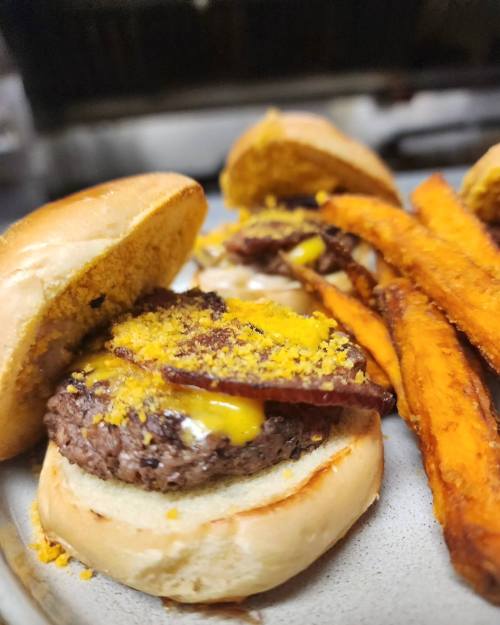 A close-up of a cheeseburger with bacon on a bun, served with sweet potato fries on the side.