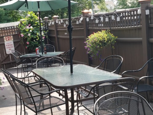 Outdoor patio with empty tables and chairs, shaded by umbrellas, surrounded by greenery and a wooden fence.