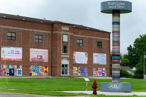A large colorful spool of thread stands in front of a brick building with vibrant murals and banners.