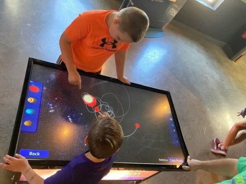 Two children interact with a touchscreen display showing a colorful solar system simulation.